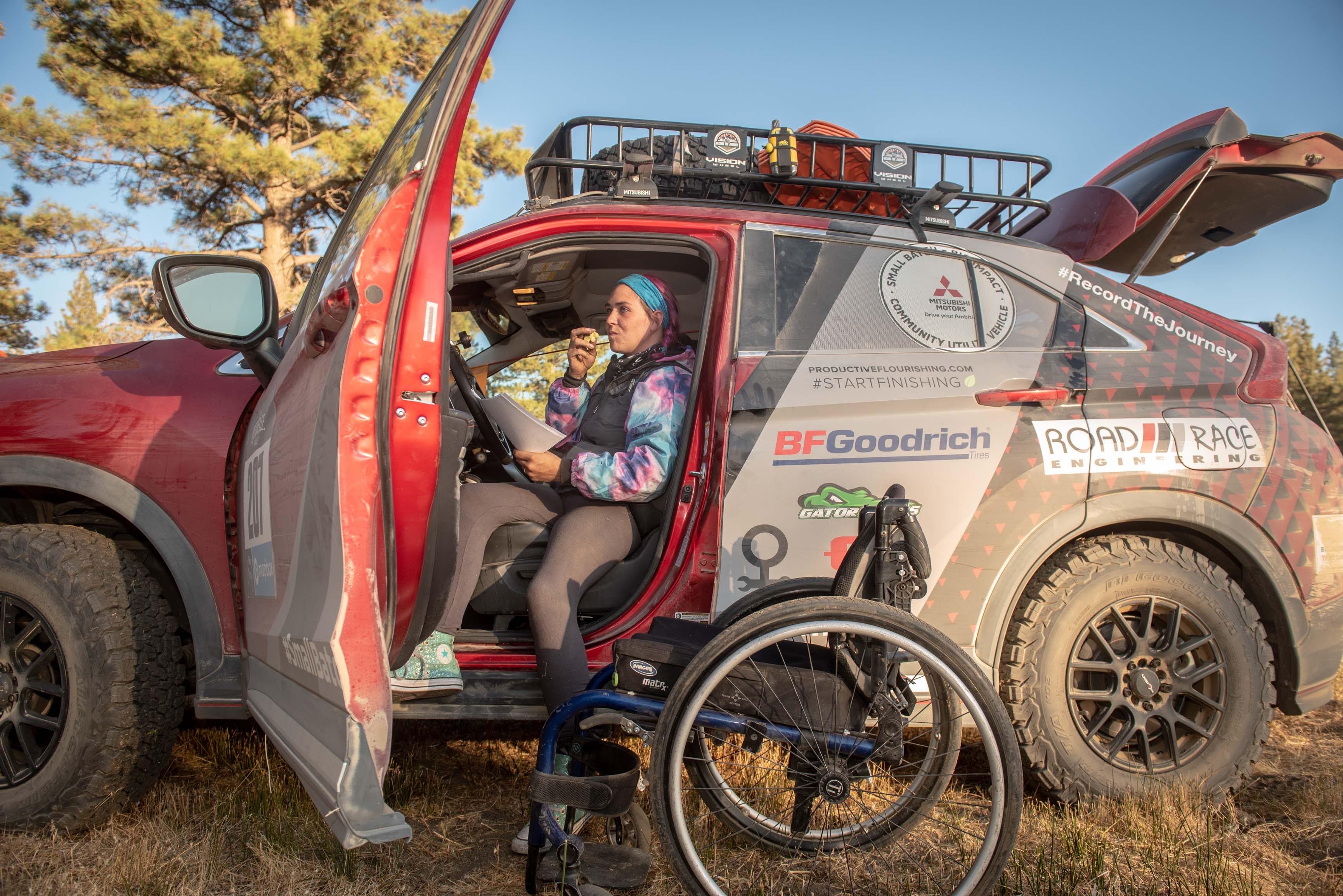 Adaptive off-road athlete Karah Behrend sits in driver seat of SUV with door open and legs hanging out the side of the SUV. Her wheelchair sits in the dirt next to the SUV