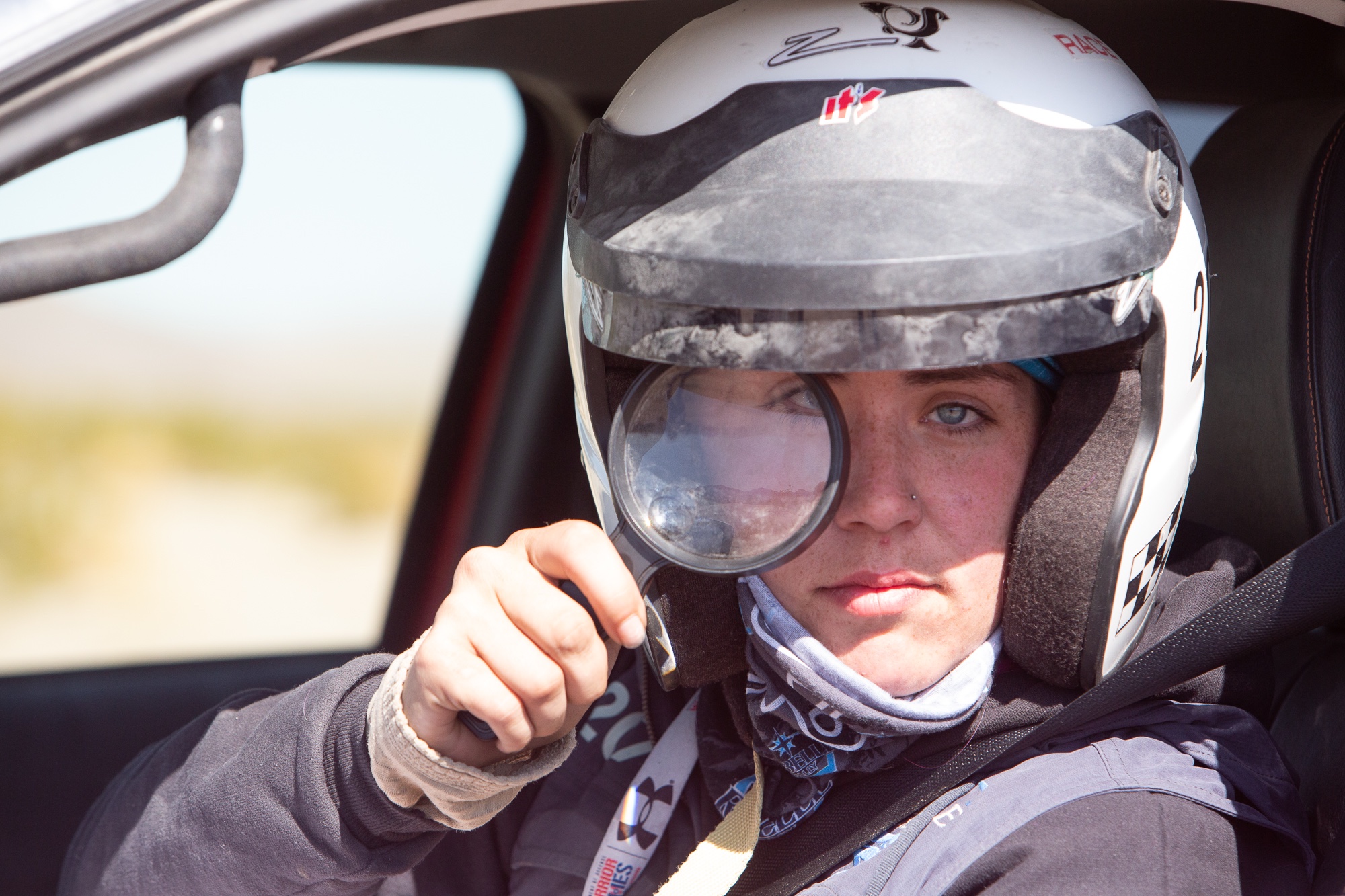 Closeup of adaptive off-road athlete Karah Behrend looking through a magnifying glass at camera while sitting in her vehicle and wearing a white helmet