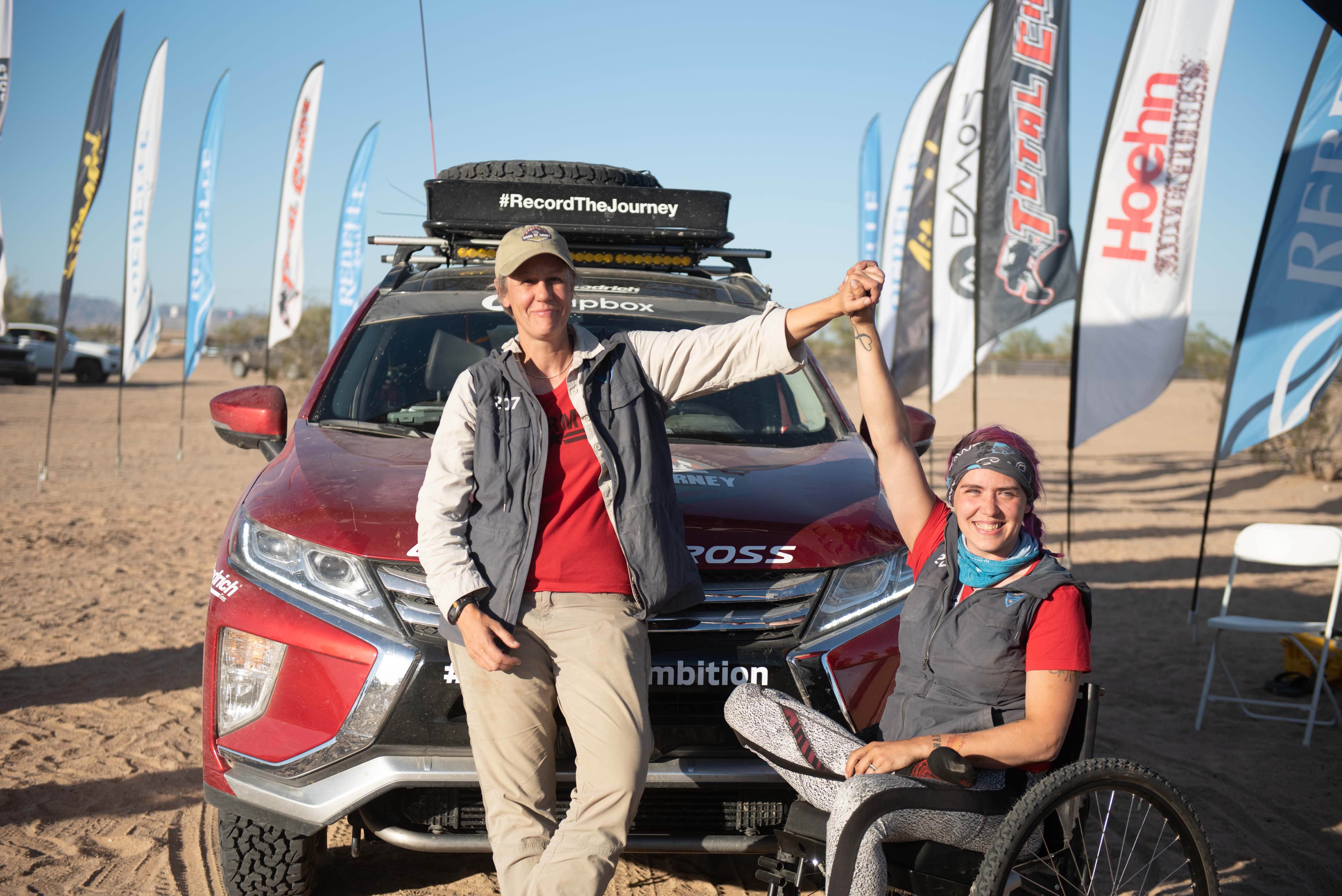 Adaptive off-road athlete Kara Behrend shown sitting in front of Mitsubishi SUV, arm raised clutching the hand of her teammate.