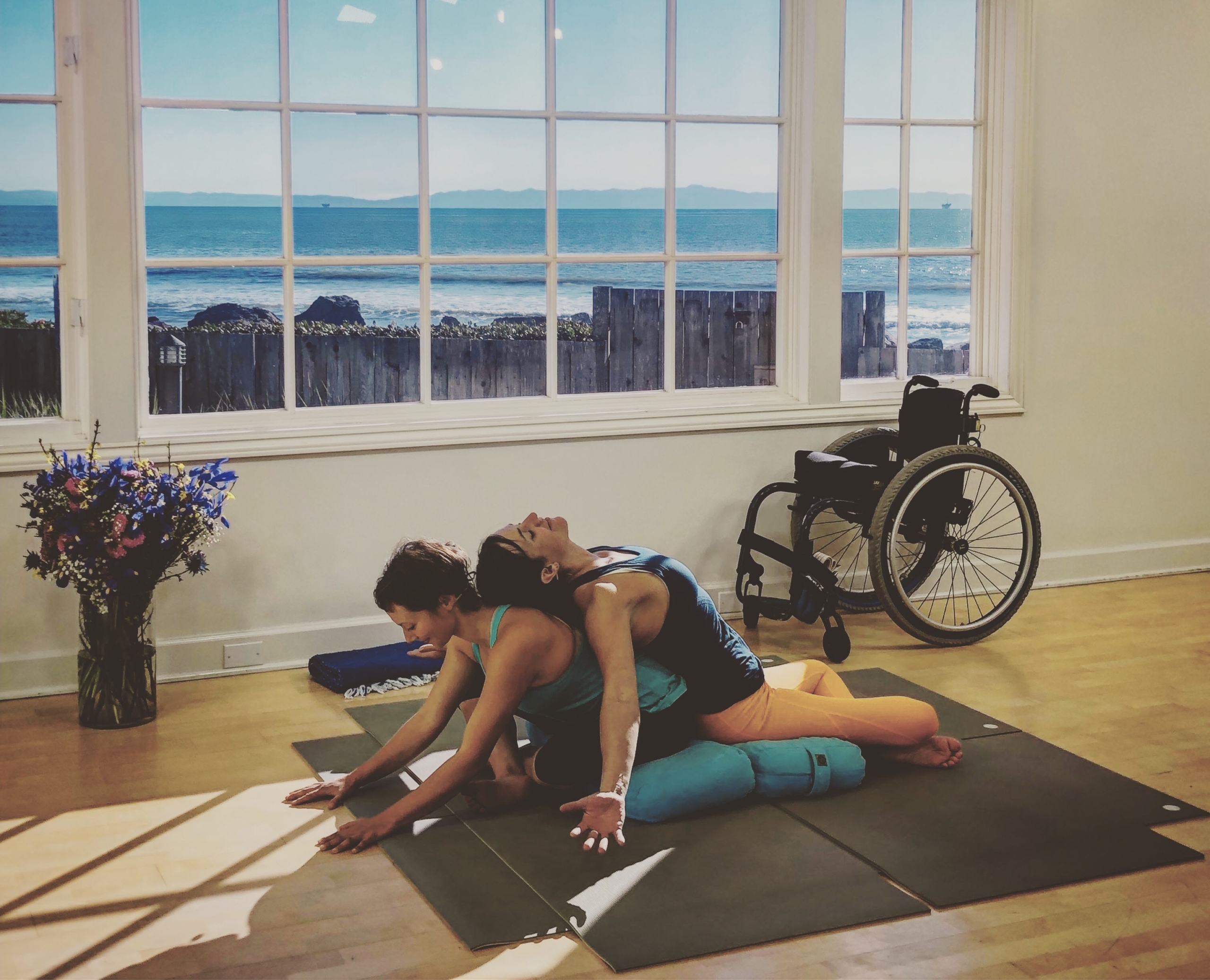 Two women in a yoga workout pose, one stretched over the others back, sitting in front of window, empty manual wheelchair in background