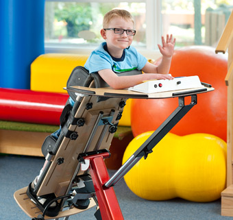 A boy standing in a Rifton Prone Stander, smiling and waving